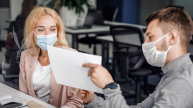 Modern ofiste sosyal uzaklık sırasında grafikler ve diyagramlarla çalışmak. Korumalı maskeli genç adam ve kadın yöneticiler Coronavirus salgını sırasında cam pano aracılığıyla belgelerle çalışırlar.