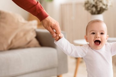 Bebek kız, babasının elini tutarak yürümeyi ve ayakta durmayı öğreniyor, baba, bebeğe evde ilk adımları atmasında yardım ediyor. Babalar Destek ve Babalık Konsepti. Kırpılmış, Seçici Odaklanma