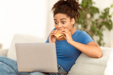 Afro-Amerikalı Kız Burger yiyor dizüstü bilgisayarla evdeki koltukta oturuyor. Aşırı yeme ve Sağlıksız Beslenme.