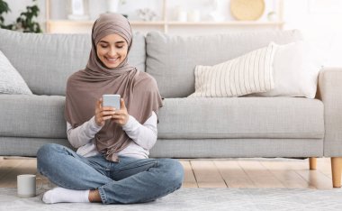 Modern Arap kız tesettürlü akıllı telefondan e-postaları kontrol ediyor, evde yerde oturuyor ve rahatlıyor, boş yer var.
