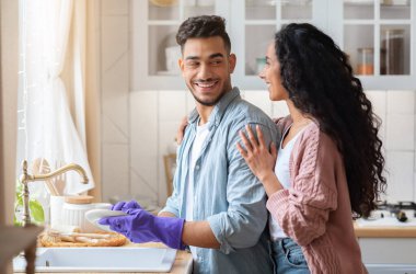 Mutfakta bulaşık yıkarken kocasını kucaklayan sevecen bir Arap kadını, ev işlerinde yardımcı olduğu için minnettar olan mutlu bir Orta Doğulu kadın. Genç Aile Yemekten sonra Tabakları Temizliyor