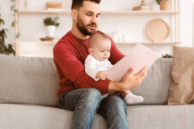 Baba, yeni doğmuş bebek için masal kitabı okuyor ve evde koltukta oturan babalığın tadını çıkarıyor. Babalar Sever, Sevimli Ebeveynlik Yaşam Tarzı Anları