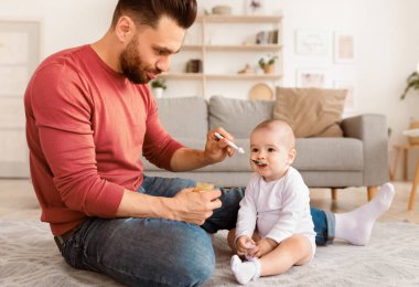 Babalık izni. Baba, evde yerde oturan kaşıktan küçük kızını besleyen çocuk. Genç baba, tatlı bebekle ilgileniyor. Babalık, Çocuk Bakımı ve Yeni Doğanlar İçin Sağlıklı Beslenme
