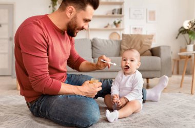 Mutlu baba kaşığı bebeğini besliyor. Babalık izni sırasında bebeğine bakıyor. Oturma odasında yerde oturuyor. Çocuk Bakımı ve Babalık kavramı