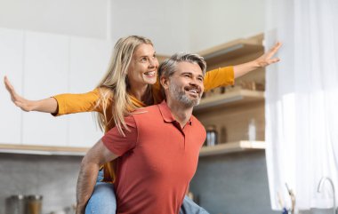 Happy pretty blonde woman piggybacking her cheerful husband, looking at copy space and smiling, middle aged couple going to cook nice meal together, having fun, kitchen interior