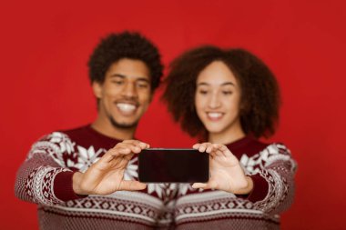 Yılbaşı şenliği ve aile için Noel özçekimi. Gülümseyen Afro-Amerikan erkek ve kadın kazaklar içinde ellerinde akıllı telefonlar ve fotoğraf çekiyorlar, boş ekranlı cihazlara odaklanıyorlar, kırmızı arka planda izole edilmişler.