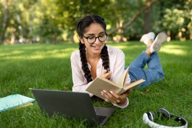 Gözlüklü mutlu Hintli kız dışarıda kitap okuyor, sınavlara hazırlanıyor ve parkta ya da üniversitede ders çalışmaktan keyif alıyor, çimlerde dizüstü bilgisayarla yatıyor.