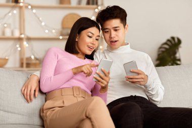 Asyalı çift cep telefonlarını kullanarak evdeki koltukta oturarak interneti tarıyor. Hafta sonu cep telefonlu aile çifti Akıllı Telefonlar İçin Uygulama Kullanıyor.