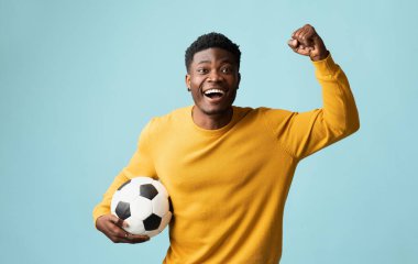 Duygusal milenyum siyahi bir adam futbol topuyla mavi stüdyo arka planında poz veriyor, Afro-Amerikan genç futbol fanatiği maça gidiyor, yumruğunu kaldırıyor, uzayı kopyalıyor. Spor konsepti