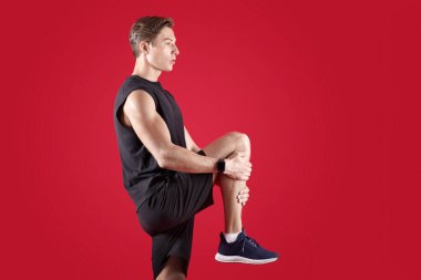 Esnek bir milenyum adamı bacağını kırmızı stüdyo arka planına doğru uzatıyor. Spor giyimli atletik genç adam antrenmandan önce ısınıyor, esneklik egzersizleri yapıyor.