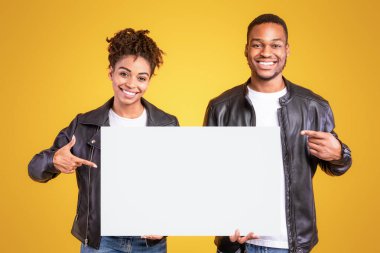 Gülümseyen Afrikalı Amerikalı çift, sarı arka planda duran bir şeyin reklamını yapmak için boş beyaz tahtayı işaret ediyorlar. Stüdyo çekimi. Reklam Afişi