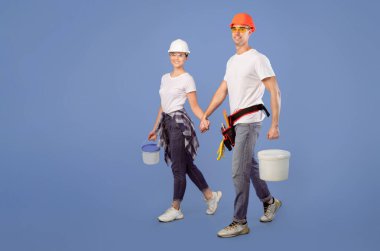 Kasketli ve koruyucu gözlüklü gülümseyen ressamlar mavi stüdyo arka planında boya kovalarıyla el ele yürüyorlar ve yenilenmeye hazırlar. Tam boy kaplama işçilerinin aile fotoğrafı.