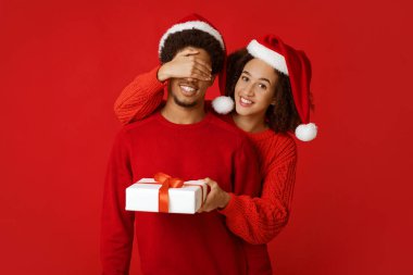 Noel için sürpriz. Gülümseyen Afrikalı genç Amerikalı Noel Baba şapkalı kadın süveter giyen adamın gözlerini kapar ve ona hediye verir, kırmızı arka planda, stüdyo çekimlerinde, kopyalama alanlarında izole edilmiş.