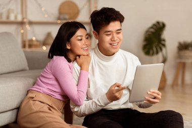 Genç Asyalı eşler, dijital tablet kullanarak evlerinde dinleniyorlar ve modern oturma odasında oturuyorlar. Hafta sonları bilgisayar kullanan Çinli bir çift. Gadget Yaşam Tarzı Konsepti