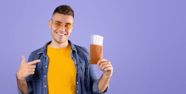 Deniz aşırı tatil konsepti. Mutlu erkek gezgin mavi stüdyo arka planında uçak biletleriyle uluslararası pasaportu işaret ediyor. Belgeleri yurtdışına seyahat için hazır olan eğlenceli turist.