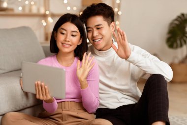 Asyalı bir çift, dijital tabletle evde oturup selam verirken video görüşmesi yapıyor. Çevrimiçi Video Konferansı için Modern Yaşam Tarzı, Uzaktan İletişim ve Uygulama