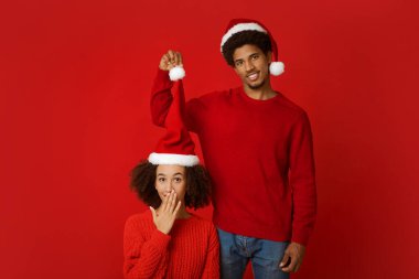 Noel 'de ya da yeni yıl partisinde iyi eğlenceler. Kırmızı arka planda izole edilmiş komik genç Afrikalı Amerikalı çift. Adam Noel Baba şapkasını gülümseyen kadının başında tutuyor. Kadın ağzını eliyle kapatıyor.
