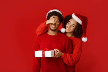 Ruh eşi için hoş bir sürpriz ve Yılbaşı ve Xmas 'ta hediye. Noel Baba şapkalı mutlu Afrikalı Amerikalı kadın süveter giyen adamın gözlerini kapar ve hediye verir, kırmızı arka planda izole edilmiş, stüdyo çekimi