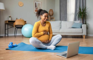 Happy young pregnant african american woman with big belly watching video workout lesson on laptop, training in living room interior. Motherhood, lady enjoy pregnancy at home, sports and coach remote