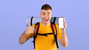 Çevrimiçi seyahat rezervasyonu. Heyecanlı genç adam cep telefonunu boş ekranla gösteriyor ve mavi stüdyonun arka planında işaret veriyor. Mobil uygulama veya web sitesi tasarımı için model