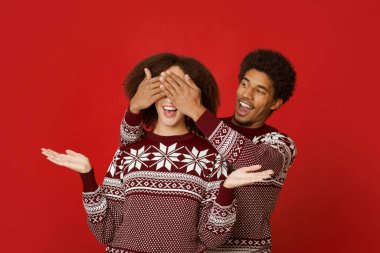 Xmas için en iyi hediye ve hoş bir sürpriz. Mutlu Afro-Amerikan erkeği, kadının arkasında duruyor ve gözlerini kapatıyor. Süveter giymiş, ağzı açık, kollarını yana açmış, kırmızı arka planda şaşırmış genç bir bayan.
