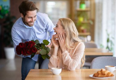 Özel bir olayı kutluyoruz. Romantik genç adam, kafedeki heyecanlı kız arkadaşına bir buket kırmızı gül sunuyor. Lokantada Sevgililer Günü için çiçekli güzel bir milenyum çifti.