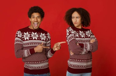 Çift olarak ilişki ve tatilde şaka. Neşeli Afrikalı Amerikalı adam, ağzı açık, parmaklarını gücenmiş kadını işaret ediyor, kollarını göğsünde kavuşturuyor, kırmızı arka planda izole edilmiş, stüdyo çekimi.