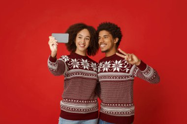 Kırmızı Noel elbiseli iki aşık selfie çekiyorlar. Genç Afro-Amerikan erkek ve kadın akıllı telefon kamerasına bakıyor. Erkek, kırmızı arka planda izole edilmiş bir şekilde barış işareti yapıyor.