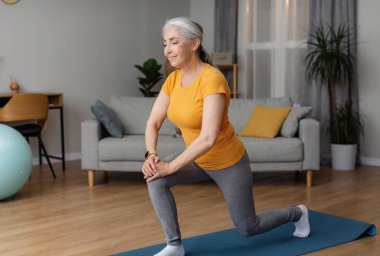 Evde formda kalan, hamleler yapan, yoga minderinde egzersiz yapan ve gülümseyen sportif son sınıf öğrencisi. Emeklilikte sağlıklı kalmak