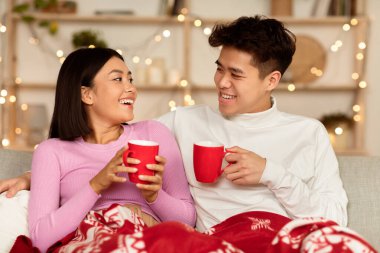 Neşeli Çinli çift, Noel arifesini evde rahat bir oturma odasında battaniyeyle kutlayarak kahve içiyorlar. Mutlu Kış Tatilleri, Yeni Yıl Aile Eğlencesi Konsepti
