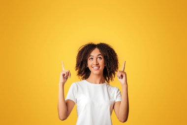 Terfi, indirim, önemli bilgiler ve metin için ilan. Gülümseyen binlerce yıllık beyaz tişörtlü Afro-Amerikan kadın sarı arka planda izole edilmiş, stüdyo çekimlerinde boş yere parmak gösteriyor.
