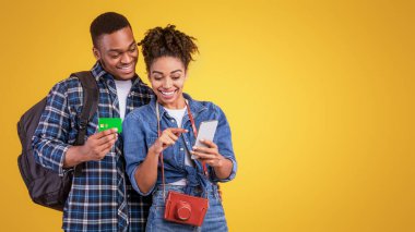 Smartphone ve Credit Card 'ı kullanarak mutlu siyah çift seyahat biletleri Sarı Stüdyo Arkaplanının üzerinde duruyor. Yurtdışında Mobil Bankacılık, Seyahat Bütçesi ve Finans.