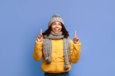 Yukarı bak. Kış ceketli Afrikalı Amerikalı Kadın Şapka ve Atkı Parmakla Gösterip Mor Stüdyo Arkaplanının Üzerinde Duran Bir Şey Öneriyor. Kış Teklif Reklam SancağıName