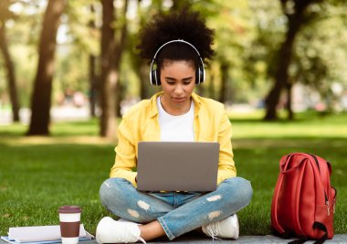 E-Öğrenme Konsepti. Afro-Amerikalı Öğrenci Kız Laptop 'ta Ders İzliyor Uzaktan kumandalı Kulaklıkla Dışarıda Park' ta Oturuyor. Modern Uzaklık Eğitimi