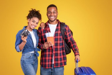 Afrika kökenli Amerikalı eşler seyahat biletleri ve bavullu pozlar Sarı Stüdyo arka planında tatil için hazırlar, kameraya gülümsüyorlar. Seyahat ve Turizm, Tatil Yolculuğu Konsepti