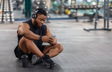 Beden eğitiminde yerde oturup müzik dinleyen, cep telefonu, kablosuz kulaklık kullanan, fotokopi çeken yorgun zenci bir vücut geliştirici. Kulaklıklı Afro-Amerikalı genç adam elinde akıllı telefon tutuyor.