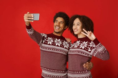 Noel veya yeni yıl partisinde mutlu bayramlar ve selfie 'ler. Gülen Afrikalı genç bayan ve adam akıllı telefona bakıyor. Kırmızı arka planda izole edilmiş bir şekilde barış işareti gösteren bir kadın.