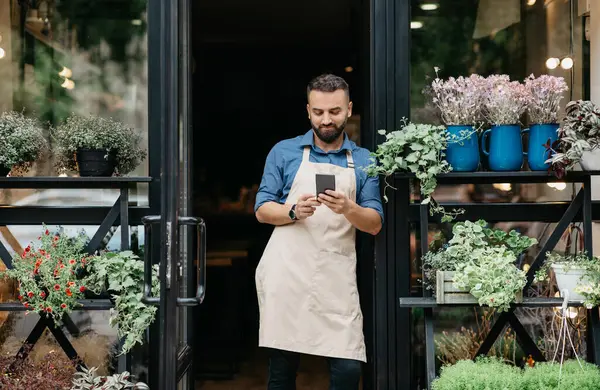 İnternette çiçekler hakkında blog, modern stüdyoda çiçekçilerin çalışmaları ve çiçek siparişi. Önlüklü neşeli genç sakallı Avrupalı adam farklı bitkilerle kırsal kesimlerin kapısında akıllı telefonuyla yazı yazıyor.