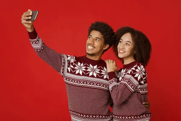 Benzer yeni yıl kıyafetleri içindeki adam ve bayan selfie için mutlu bir poz veriyorlar. Gülümseyen Afrikalı Amerikalı çift akıllı telefondan izliyor, Noel kartı için fotoğraf çekiyorlar, kırmızı arka planda izole edilmiş, stüdyoda çekiliyor.