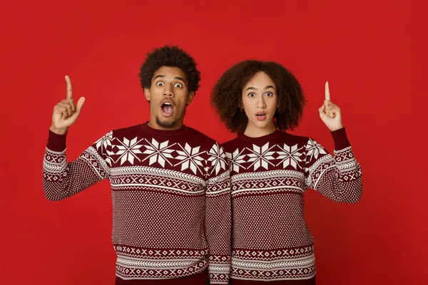 Sürpriz ve şok, doğal insan duyguları. Ağızları açık, parmakları boş, kırmızı arka planda izole edilmiş, Noel süveteri giyen Afro-Amerikalı genç adam ve bayan.