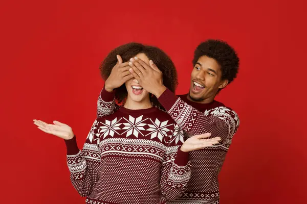 Xmas için en iyi hediye ve hoş bir sürpriz. Mutlu Afro-Amerikan erkeği, kadının arkasında duruyor ve gözlerini kapatıyor. Süveter giymiş, ağzı açık, kollarını yana açmış, kırmızı arka planda şaşırmış genç bir bayan.
