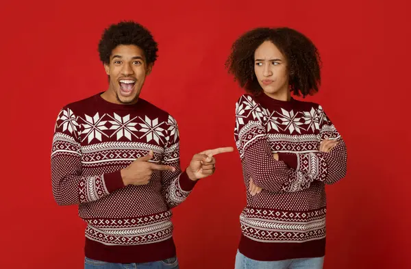 Çift olarak ilişki ve tatilde şaka. Neşeli Afrikalı Amerikalı adam, ağzı açık, parmaklarını gücenmiş kadını işaret ediyor, kollarını göğsünde kavuşturuyor, kırmızı arka planda izole edilmiş, stüdyo çekimi.
