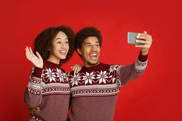 Yılbaşı ve Noel tatilleri boyunca aileye video bağlantısı. Mutlu Afro-Amerikalı adam telefonu tutuyor, kazaklı kadın el sallıyor ve akıllı telefona bakıyor, kırmızı arka planda izole edilmiş, stüdyo çekimi.