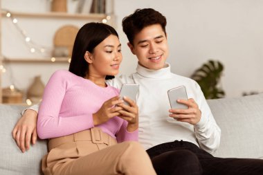 Akıllı telefonlar kullanan Asyalı çift evdeki koltukta birlikte oturarak interneti tarıyor. Çevrimiçi Sosyal Medya Ağı ve Mesajlaşma Kullanan Eşler. Cep telefonu Kavramı için Cep telefonu Uygulaması
