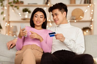 Mobil Banka Uygulaması. Zeki telefonları ve kredi kartlarıyla internetten alış veriş yapan Asyalı çift evdeki rahat oturma odasındaki koltukta otururken internetten bir şeyler seçip alıyor. Seçici Odaklanma