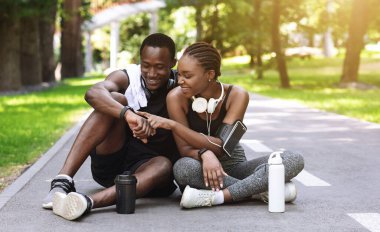 Neşeli siyah çift parkta koştuktan sonra dinleniyor, patikada otururken akıllı spor takip cihazlarını kontrol ediyor, dışarıda antrenmanların keyfini çıkarıyor, boş alan arıyor.