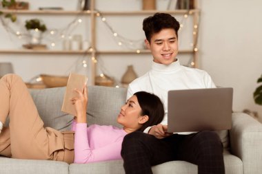 Japon eşler dizüstü bilgisayarla rahatlıyor ve kitap okuyor hafta sonları evde, oturma odasında koltukta oturuyor. Homey Evening Family Leisure, birlikte zaman geçiren çiftler.