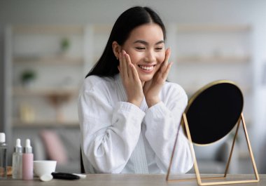 Bornozlu güzel bir Asyalı kadın yüz masajı yapıyor, yatak odasında masada oturuyor, aynaya bakıyor ve gülümsüyor, güzel Çinli kadın duştan sonra nemlendirici yüz kremi kullanıyor, boşluğu kopyalıyor.