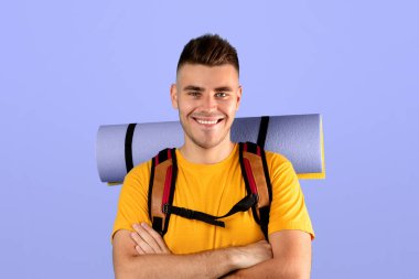 Sırt çantası ve turist minderiyle gülümseyen ve kollarını mavi stüdyo arka planında kavuşturan neşeli milenyum adamı portresi. Mutlu maceracı adam yürüyüşe ya da kamp gezisine çıkmaya hazır.