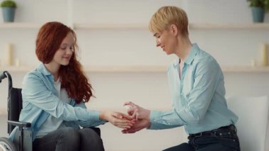 Bakıcı nazikçe tekerlekli sandalyedeki bir kızın elini tutar. Yumuşak ışıklandırma ve en az dekorla aydınlık, rahatlatıcı bir mekanda bir bağlantı anlarını paylaşıyorlar..
